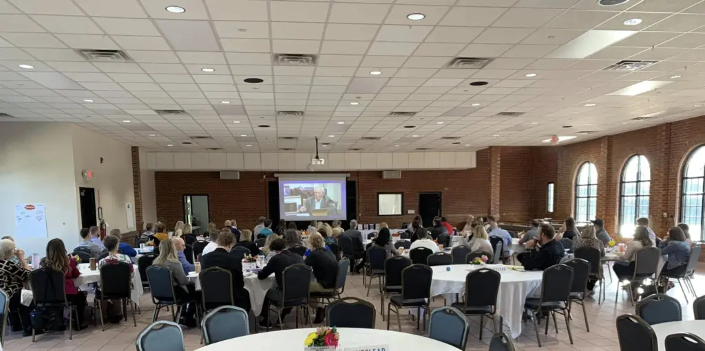 Rows of round tables fill the brick-walled Lake Storey Pavilion as attendees watch a speaker on a large projection screen