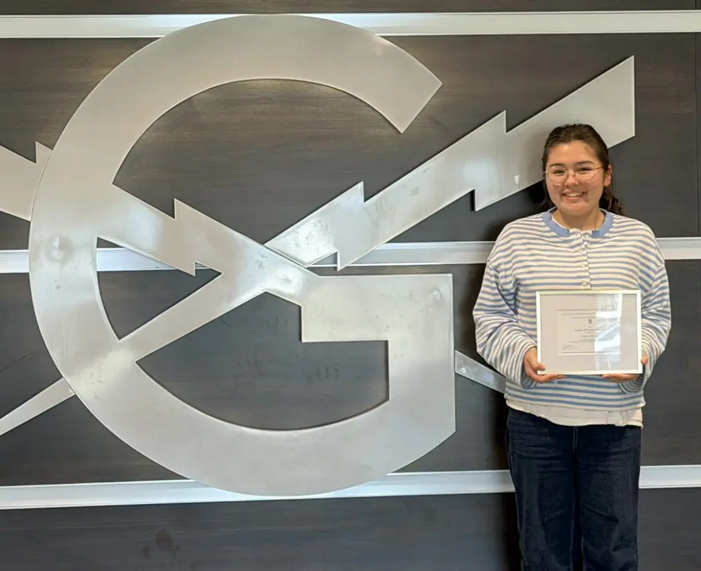 Emily Edwards smiles and holds a framed certificate while standing beside a large silver Galesburg G and lightning bolt logo on a dark wood wall