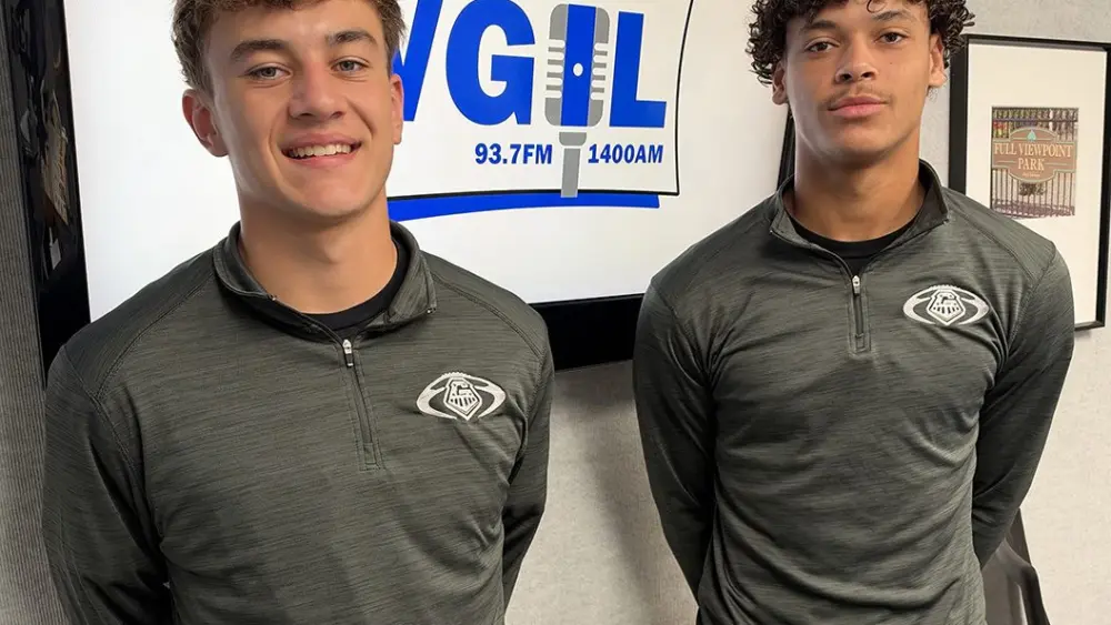 Two Galesburg High School football players in matching gray quarter-zip pullovers stand in front of a WGIL logo screen in the station’s studio
