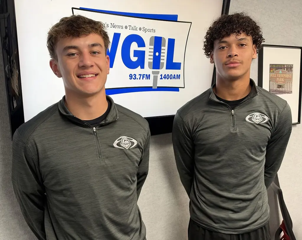 Two Galesburg High School football players in matching gray quarter-zip pullovers stand in front of a WGIL logo screen in the station’s studio