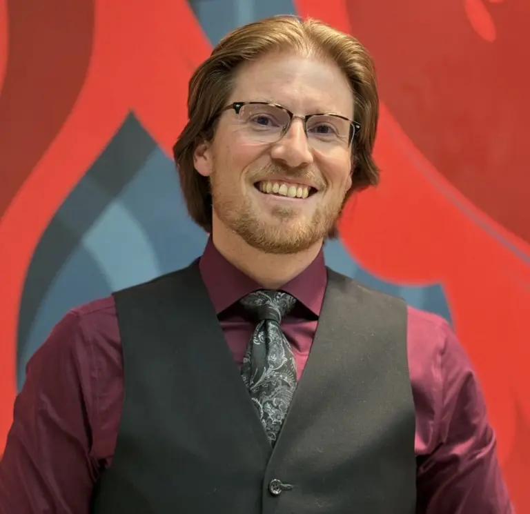 John Glasgow, smiling, wearing glasses, a dark vest, a burgundy shirt, and a patterned tie in front of a red and blue mural