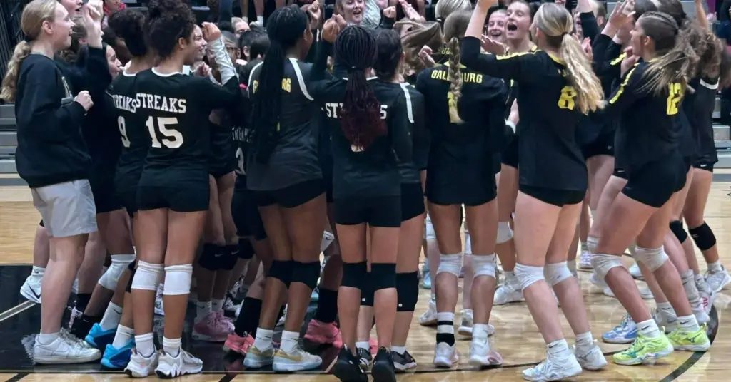 ALT TEXT: Galesburg High School volleyball players in black Streaks jerseys celebrating in a group on the gym floor