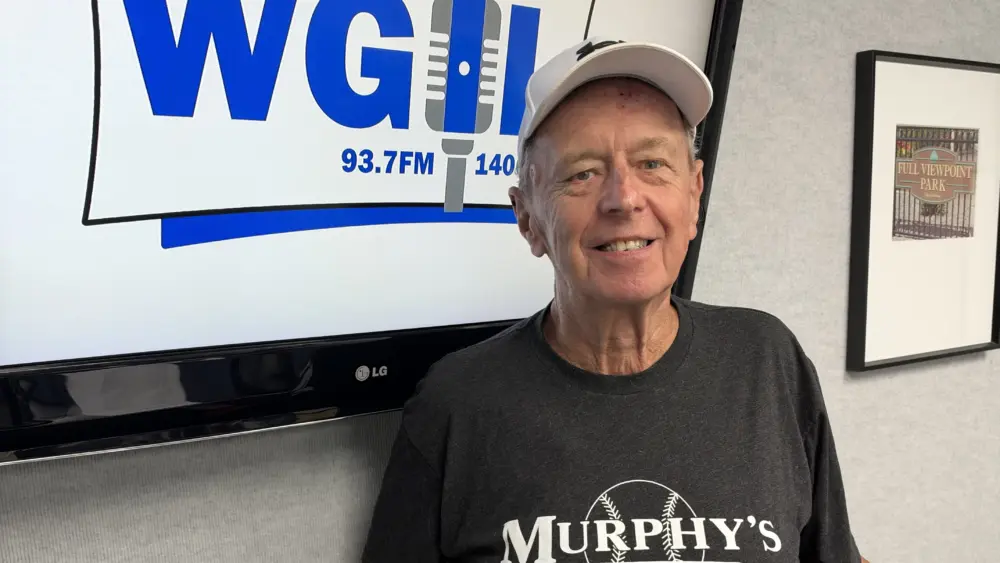 ALT TEXT: Mike Trueblood in a white cap and Murphy’s Bleachers T-shirt standing in front of a WGIL logo on a studio screen
