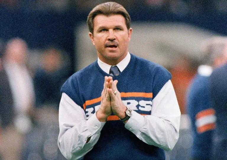 Mike Ditka in a navy Bears sweater vest and tie, palms pressed together, on the sideline