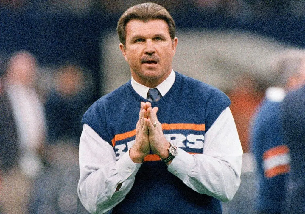 Mike Ditka in a navy Bears sweater vest and tie, palms pressed together, on the sideline