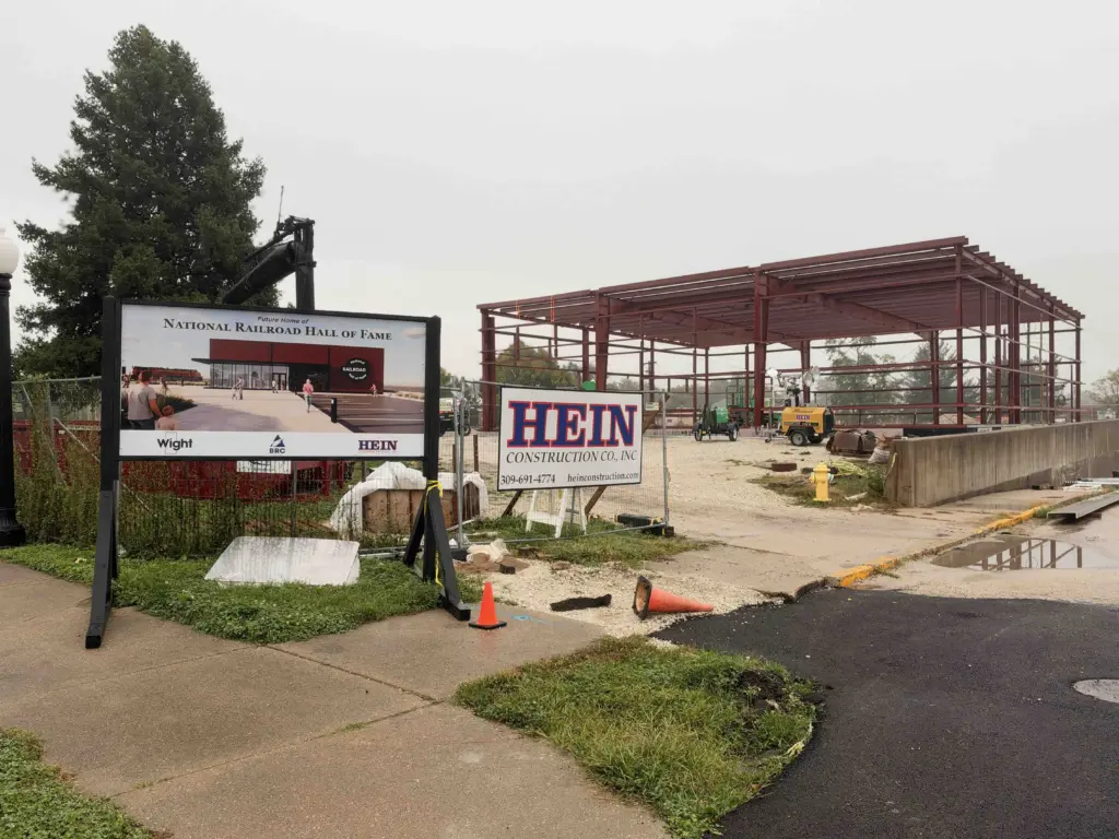 A sign showing a rendering of the National Railroad Hall of Fame stands in front of the red steel frame of the building under construction in Galesburg.