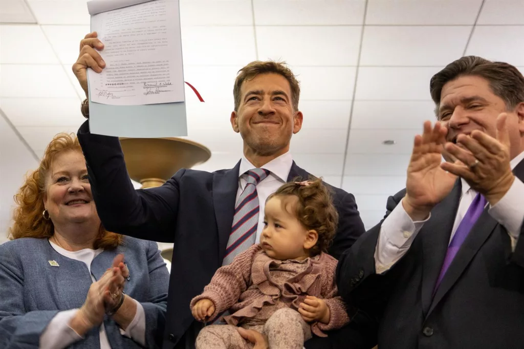 Illinois Secretary of State Alexi Giannoulias holds his daughter while brandishing a bill signed by Gov. JB Pritzker Monday that aims to discourage book bans at Illinois libraries. Bill sponsor Sen. Laura Murphy, D-Des Plaines, is pictured at left and Pritzker is pictured at right. (Capitol News Illinois photo by Andrew Adams)