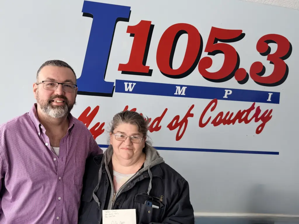 Station Manager Paul Jones stands in front of the I105.3 logo with Bingo Winner Meleana Baker who is holding her prize.