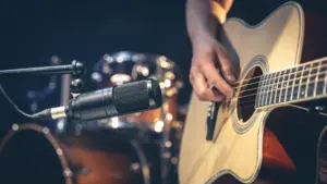 Male musician playing acoustic guitar behind microphone in recording studio.