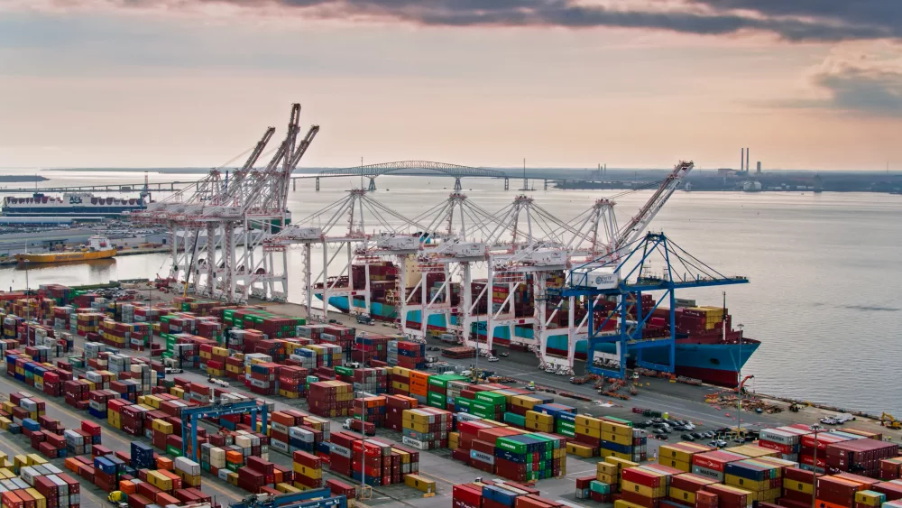 Aerial shot of the Port of Baltimore at sunset, looking across and intermodal container yard towards a ship being loaded by cranes. In the distance, the Key Bridge stretches across the river.