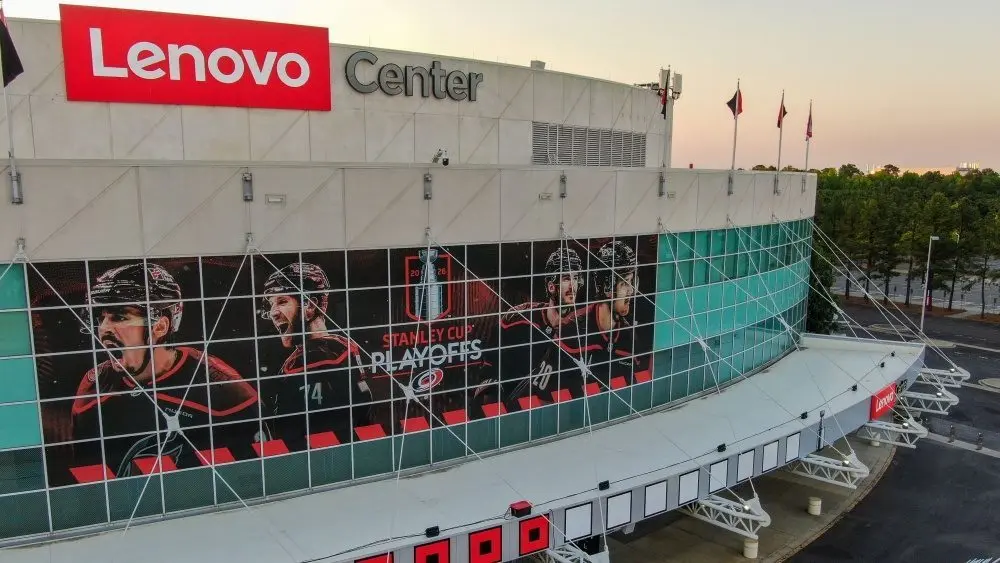 Aerial 4K drone changeable captures a high-angle twilight presumption of the Lenovo Center arena^ location of the NHL's Carolina Hurricanes. The debased aureate hr prima rakes crossed the modern facade^ highlighting the reflective teal solid panels of the Carolina Hurricanes location venue earlier they look the Montreal Canadiens. The changeable documents the wide^ geometric infrastructure layout of the bare parking lots^ main entranceway gates^ and surrounding roadway logistics nether a cleanable sunset gradient sky. Raleigh^ North Carolina^ USA - May 19^ 2026