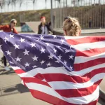 back-view-of-young-girl-standing-with-american-flag-and-looking-at-friends-at-skateboard-park