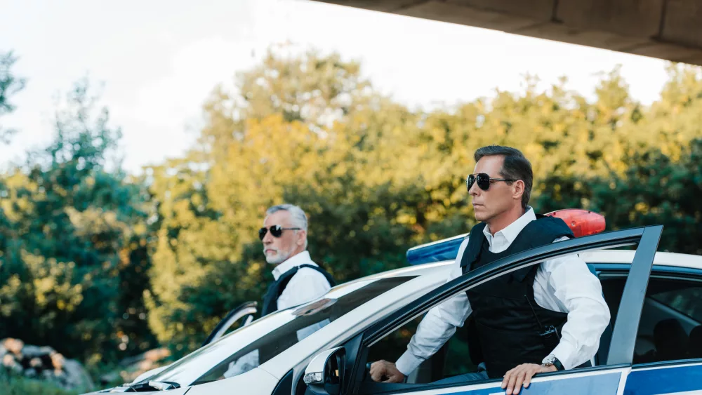 selective-focus-of-mature-policeman-in-sunglasses-and-bulletproof-vests-standing-near-car-at-city