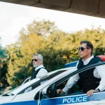 selective-focus-of-mature-policeman-in-sunglasses-and-bulletproof-vests-standing-near-car-at-city