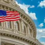 The star spangled banner American flag flies proudly in front of the US capitol building in Washington DC with blue cloudy sky background