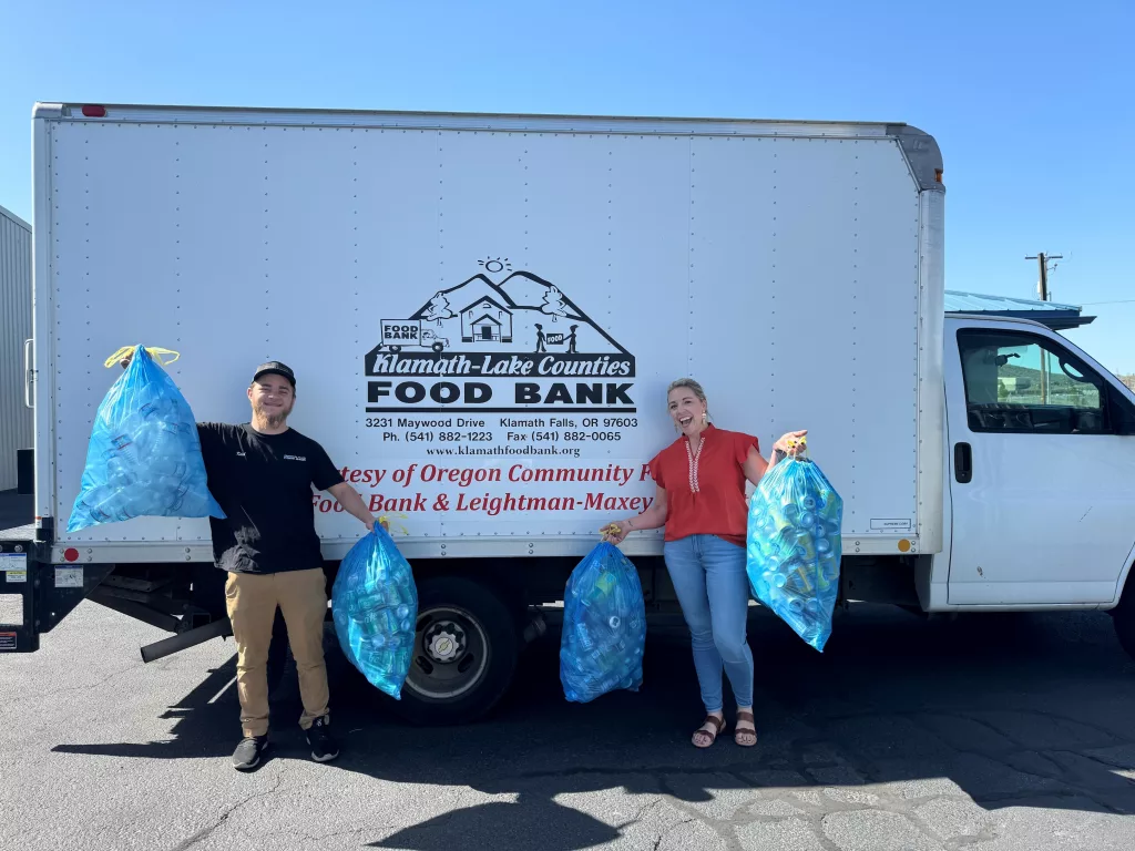 Two people hold plastics bags full of cans next to a Klamath-Lake Counties Food Bank truck