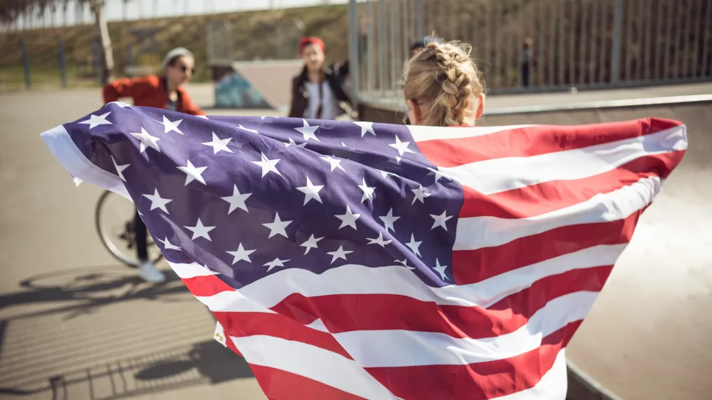 back-view-of-young-girl-standing-with-american-flag-and-looking-at-friends-at-skateboard-park
