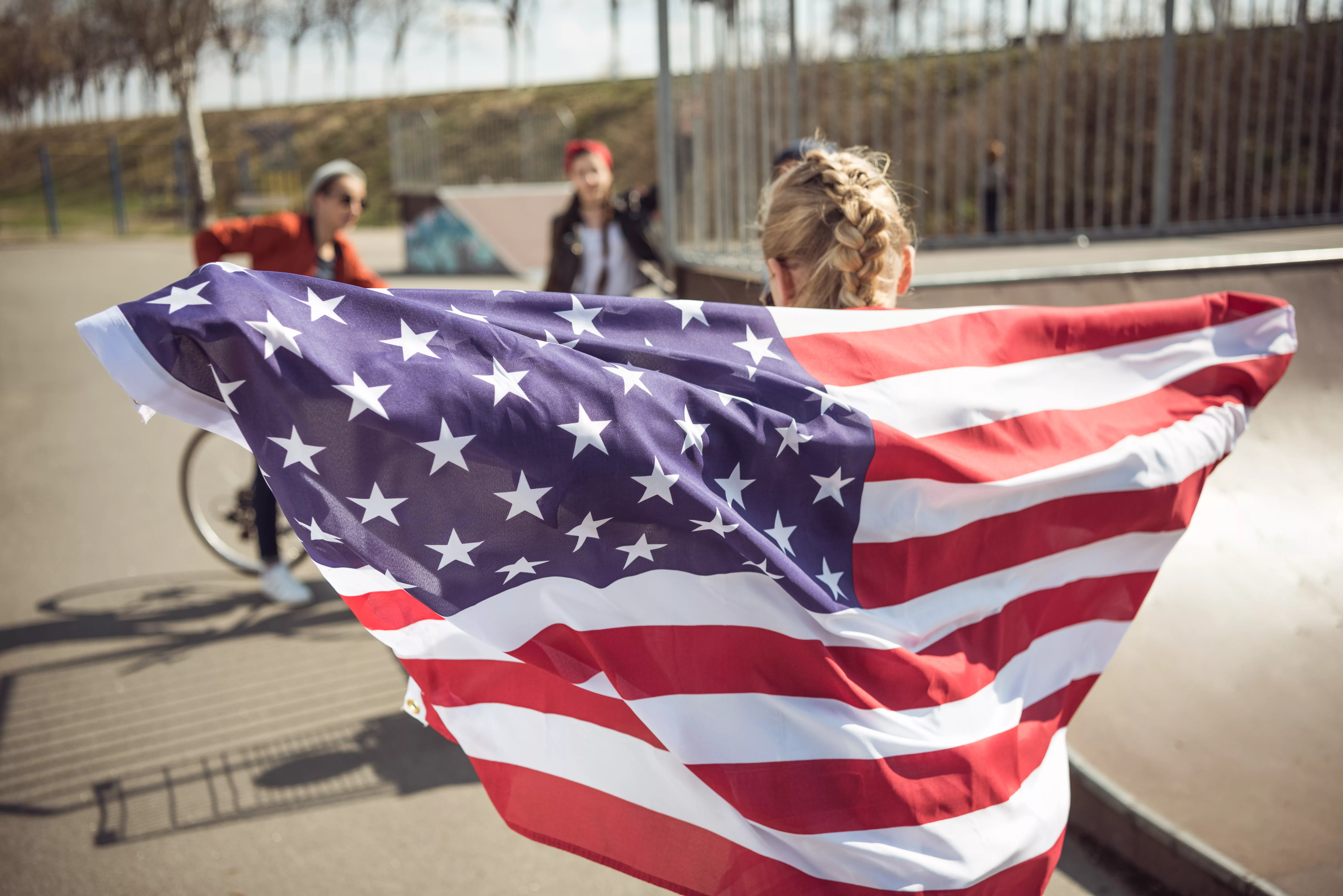 back-view-of-young-girl-standing-with-american-flag-and-looking-at-friends-at-skateboard-park