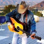 Dwight Yoakam holding a guitar on a roof top with mountains in the back ground.