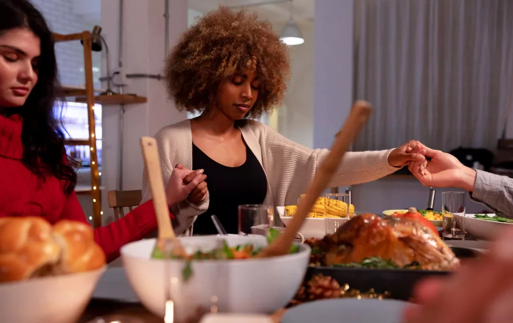 front-view-of-a-young-adult-mixed-race-woman-and-a-young-adult-caucasian-woman-sitting-around-a-table-holding-hands-with-friends-saying-grace-before-eating-thanksgiving-dinner-at-home-together-with-fr