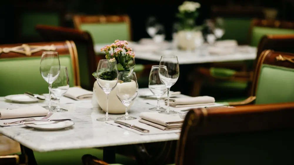 horizontal-shot-of-square-table-served-with-empty-glasses-napkins-plates-forks-and-knives-with-no-dish-green-chairs-around-cozy-restaurant-or-cafe