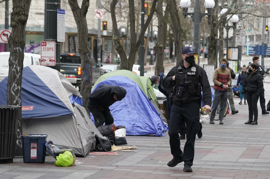 FILE - A Seattle police officer walks past tents used by people experiencing homelessness, March 11, 2022, during the clearing and removal an encampment in Westlake Park in downtown Seattle. Five major U.S. cities, including Seattle, and the state of California will receive federal help to get unsheltered residents into permanent housing under a new plan announced Thursday, May 18, 2023, as part of the Biden administration's larger goal to reduce homelessness 25% by 2025. (AP Photo/Ted S. Warren, File)