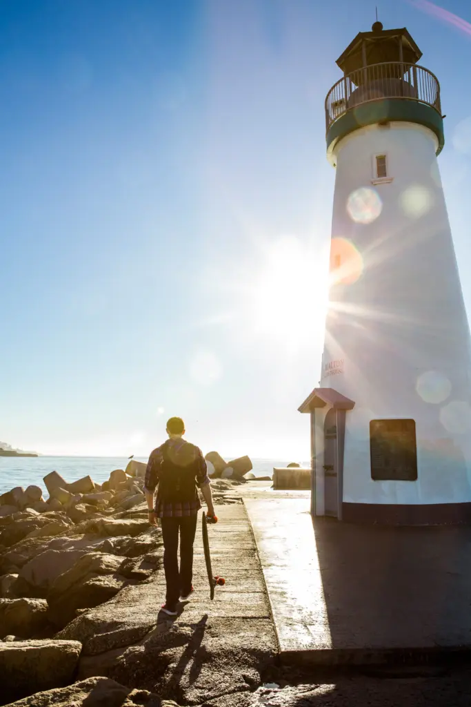 young-man-holding-skateboard-walking-beside-lighthouse-rear-view
