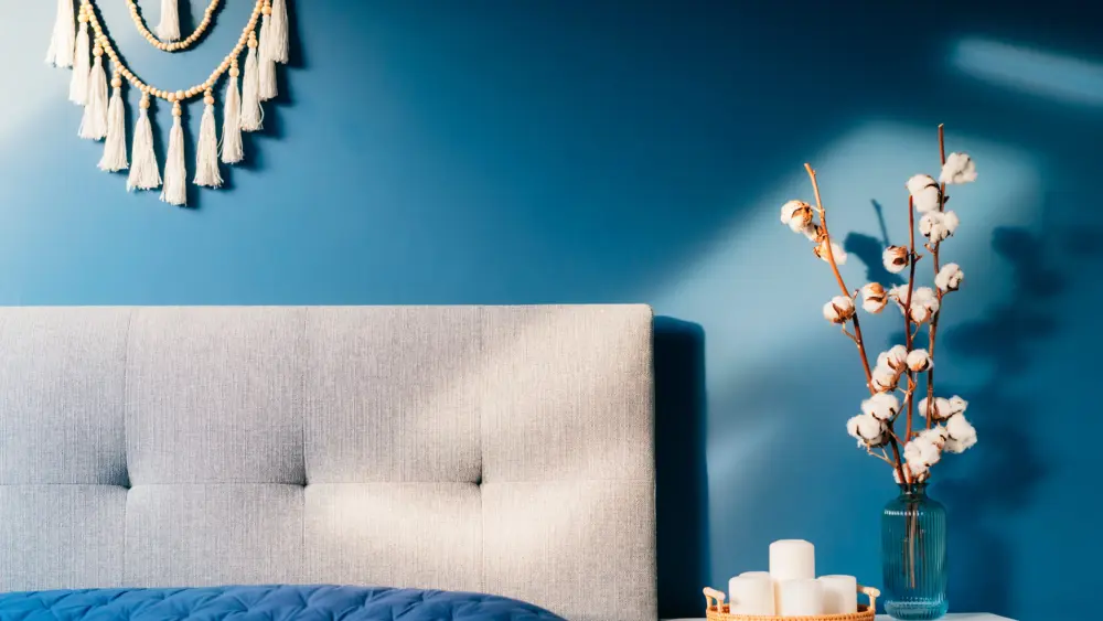 stylish-modern-bedroom-in-dark-colors-cozy-interior-with-navy-blue-walls-home-decor-bed-with-gray-fabric-headboard-blue-blanket-bedside-table-vase-with-natural-cotton-flowers-candles-on-tray