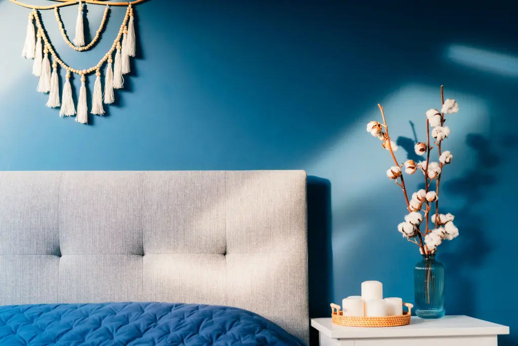 stylish-modern-bedroom-in-dark-colors-cozy-interior-with-navy-blue-walls-home-decor-bed-with-gray-fabric-headboard-blue-blanket-bedside-table-vase-with-natural-cotton-flowers-candles-on-tray