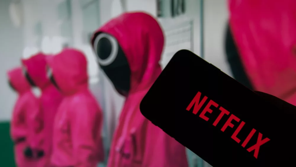 Netflix logo on the display of a smartphone in front of a television with the new series "Squid Game" ( focus on Netflix logo )