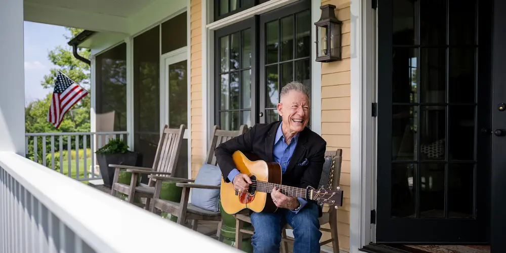 Singer-songwriter Lyle Lovett sits on his porch playing a guitar.