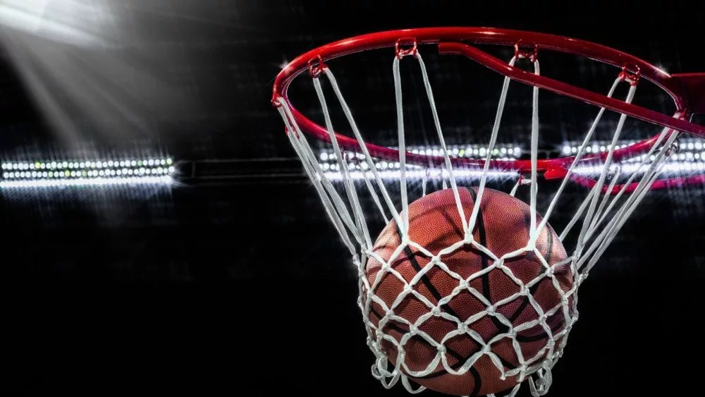 Looking up at an orange basketball falling through the rim and a white nylon net. With the arena lights in the background.