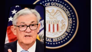 Federal Reserve (Fed) Chair Jerome Powell is visible on smartphone screen. In the background^ an inflation rate chart is displayed.