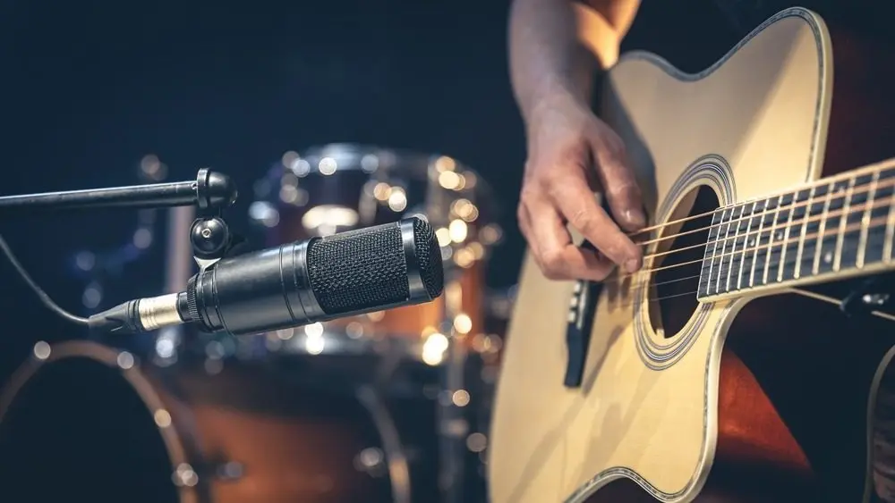 Male musician playing acoustic guitar behind microphone in recording studio.