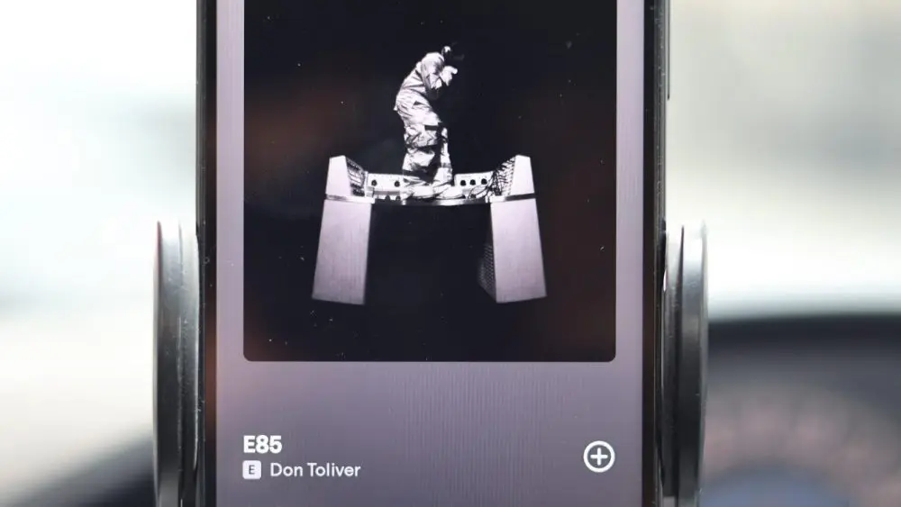 This image shows a smartphone mounted in a car's phone holder^ displaying the Spotify music player. The phone is playing a song titled "E85" by artist Don Toliver^ which is the opening track of his 2026 album^ OCTANE.