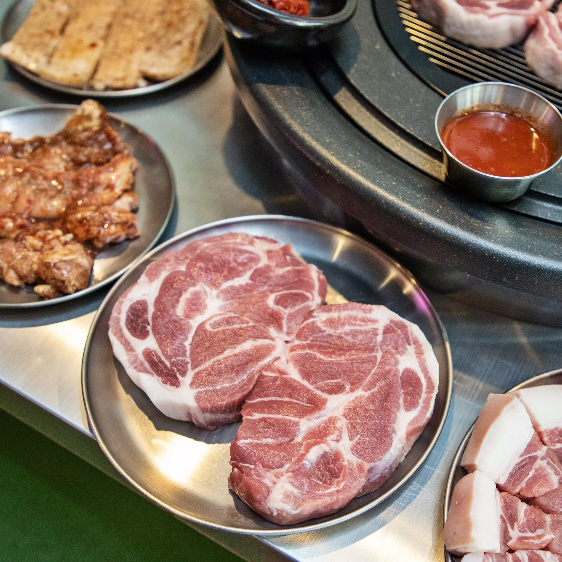 Plates with raw pork neck and other cuts of meat ready for grilling, with sauce and seasonings on the side.