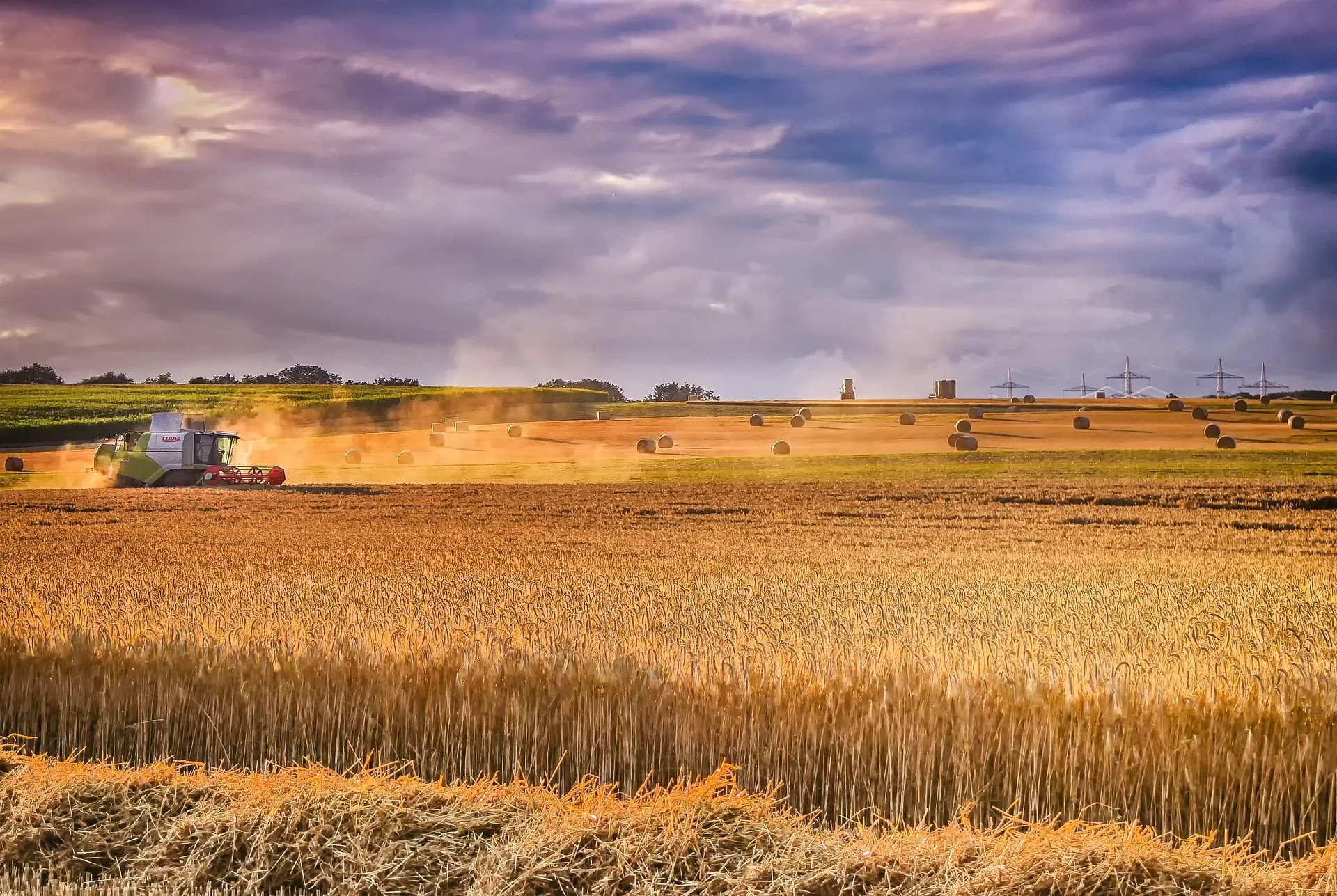 Combine harvester working in a field with hay bales and wind turbines in the background.