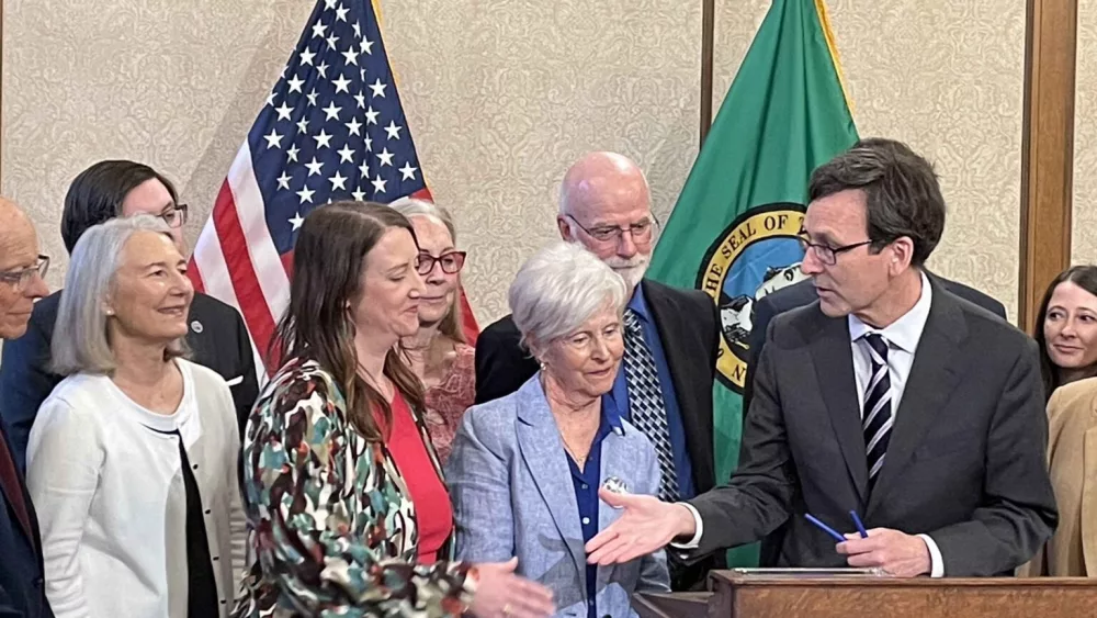 Gov. Bob Ferguson, at podium, goes to shake hands with state Sen. Noel Frame, D-Seattle, at the signing of a bill to make clergy mandatory reporters of child abuse and neglect, on May 2, 2025 in Olympia. At center is Mary Dispenza, a founding member of the Catholic Accountability Project. (Photo by Jerry Cornfield/Washington State Standard)