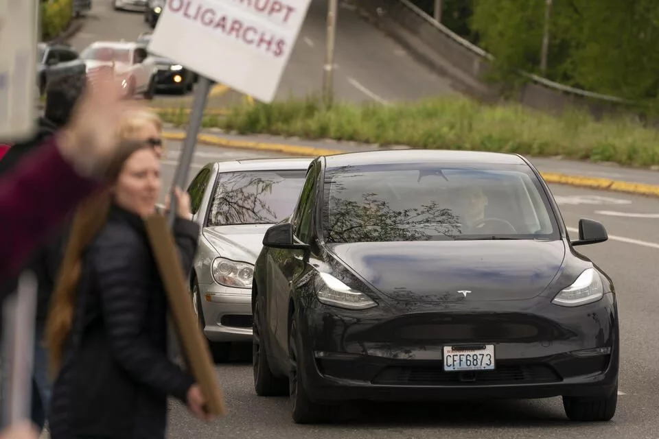 A Tesla passes by demonstrators during a Tesla protest at University Village in Seattle, Washington on May 3, 2025. Tesla protests have had a major impact on the company and its sales in the Seattle area. (David Ryder for Cascade PBS)