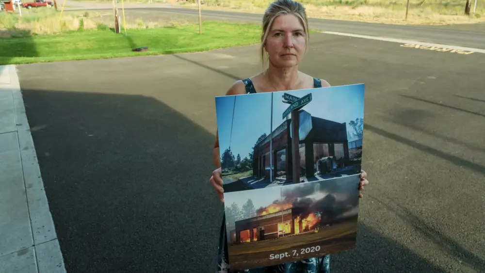 Chandelle Frick, city clerk for the town of Malden and executive director of the Pine Creek Community Long-Term Recovery Organization, stands outside the Malden Town Hall on July 31 holding photos of the town’s old post office, which was located a block away behind her, burning in the wildfire that swept through the town in 2020. The Hall is now located in a new building that also houses a post office and fire department, but other areas of the town have been slow to recover. (Ted S. Warren for Cascade PBS)
