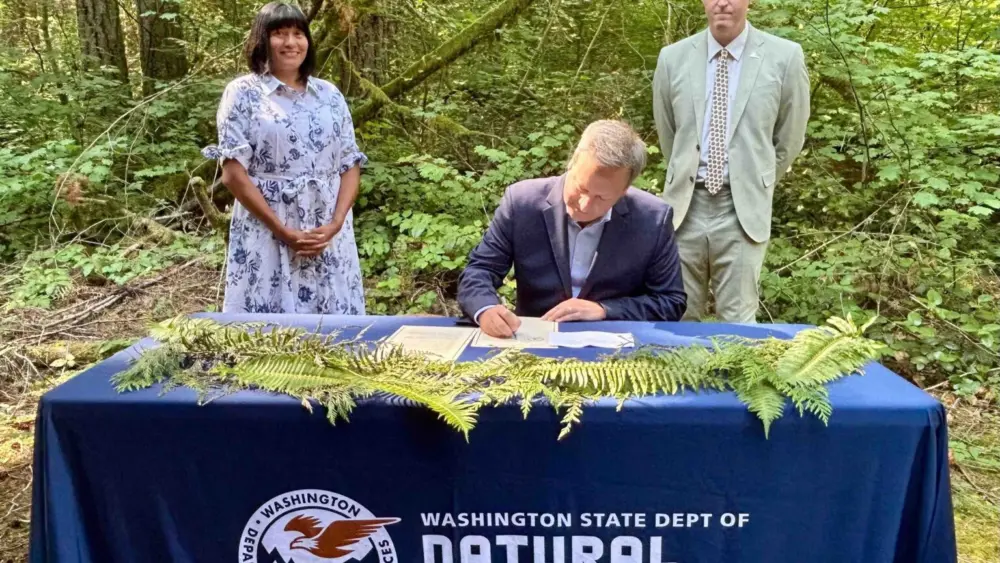 Washington Public Lands Commissioner Dave Upthegrove signs a directive ordering the conservation of about 77,000 acres of older forests on state logging lands, on Aug. 26, 2025, at the High Point Trailhead on Tiger Mountain. Behind him are Washington Conservation Action CEO Alyssa Macy, left, and Thurston County Commissioner Tye Menser, right. (Photo by Emily Fitzgerald/Washington State Standard)