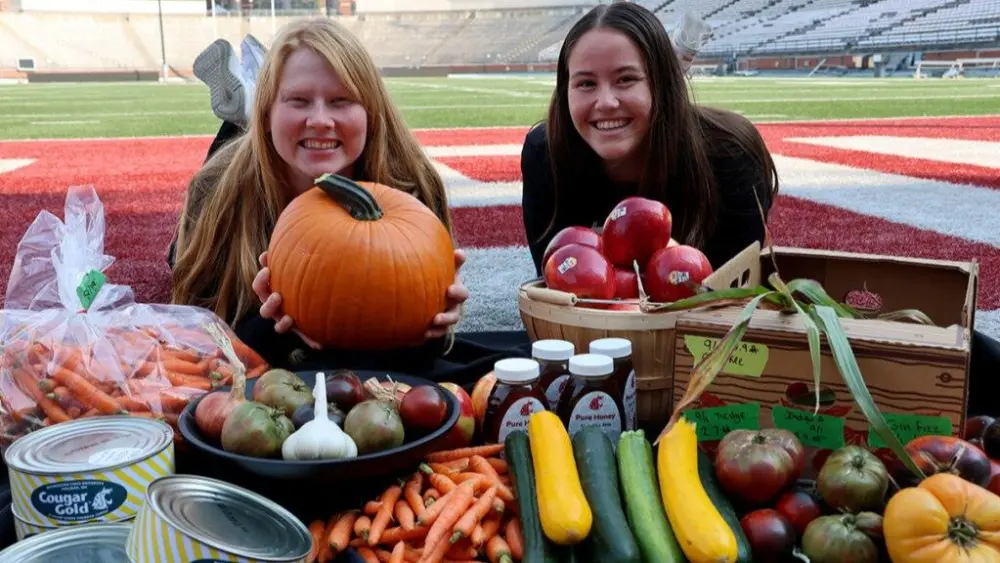 Jessica Lovell and Jessica Schneider, interns with WSU's Performance Nutrition program, show a selection of WSU-grown foods and products appearing at the upcoming Apple Cup Farmer's Market, Sept. 19. Game weekend visitors can sample iconic WSU tastes, including the Cosmic Crisp® apple, WSU-raised honey, organic produce, cheese, and more (photo by Seth Truscott/WSU).
