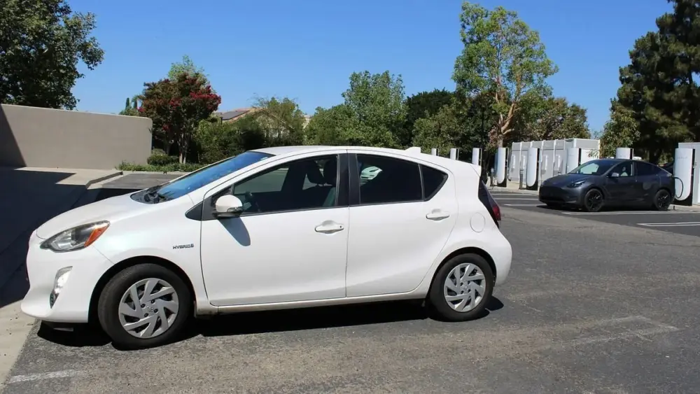 A white Toyota Prius C, a hybrid, is parked near an electric vehicle charging at a Tesla station. Dave Mason / The Center Square