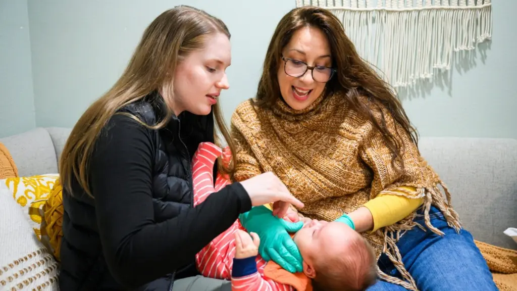 Lactation consultant Beth Waters, right, checks up on 3-month-old Reuel as he is held by his mother Rebekah Wong, left, at Andaluz Waterbirth Center in Portland, Ore., on Friday, November 14, 2025. Waters and Wong connected while Wong was looking for a way to exclusively breastfeed Reuel. (Photo by Molly J. Smith/Investigate West)