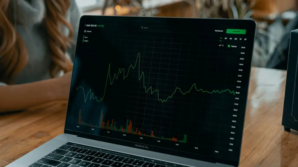A laptop screen shows a fluctuating stock market chart with trading volume bars. Photo: Tech Daily / Unsplash