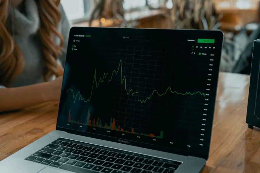 A laptop screen shows a fluctuating stock market chart with trading volume bars. Photo: Tech Daily / Unsplash