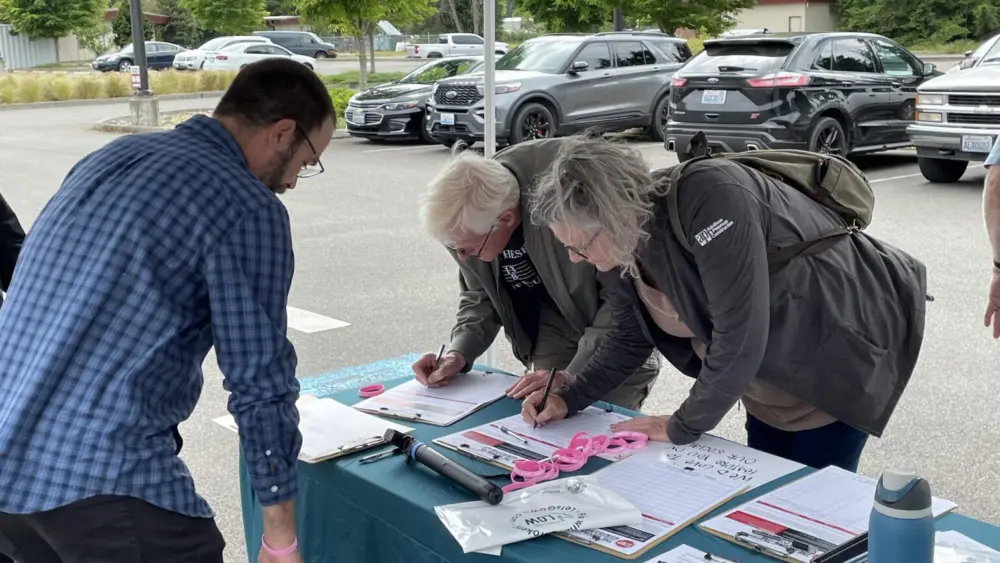 Josh Johnson (left) of Let’s Go Washington looks on as two more people sign petitions for a proposed ballot measure to repeal Washington’s new income tax on high earners. These signatures were gathered June 3, 2026 in Tumwater. (Photo by Jerry Cornfield / Washington State Standard)
