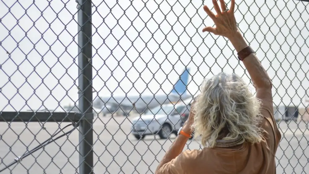 Maureen Newton waves as she uses binoculars to watch U.S. Immigration and Customs Enforcement detainees board a plane on Aug. 11, 2026, at the Magic Valley Regional Airport. (Photo by Laura Guido/Idaho Capital Sun)