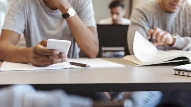 midsection-of-male-student-using-smart-phone-while-sitting-at-desk-in-classroom