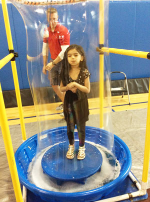 Kindergartener Aliyah Ahmad lifts a bubble around herself as part of the Kidz Science Safari while Physical Education teacher, Jeremy Boland, monitors the hands-on experiment.